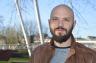 Osvaldo Bogado Pascottini with a full beard stands outdoors on a sunny day. He is wearing a brown leather jacket over a grey and black sweater. Behind him are blurred metal structures, leafless tree branches, and a bridge, suggesting a park or urban setting.